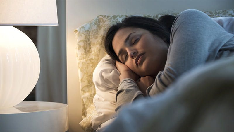An Hispanic/Latina woman illuminated by a lamp is sleeping in bed. An Hispanic/Latina woman illuminated by a lamp is sleeping in bed.