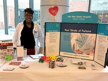 una mujer que representa a Stamp Out Stroke (Erradicar Ataques o Derrames Cerebrales) está de pie detrás de una mesa con un tríptico y otros materiales informativos del Centro de Ataque o Derrame Cerebral del Hospital Yale New Haven